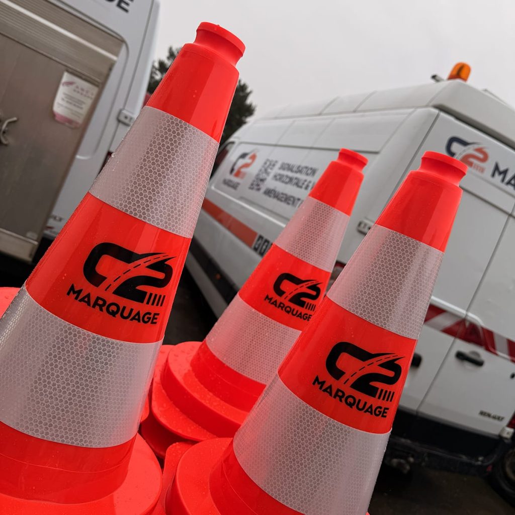 Trois cônes de signalisation orange C2 MARQUAGE en premier plan devant deux fourgons d'intervention C2 MARQUAGE sur un chantier